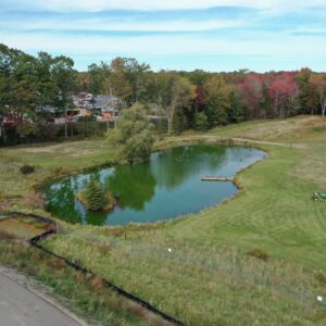 A large grassy area with a pond in the center, the pond has ducks on it and a dock along the shore. The grassy area has two riding lawnmowers parked near each other.