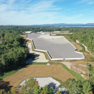 An aerial view of the Copart construction site while gravel distribution is in progress. In the foreground there are many pine trees and dense forest surrounding the worksite, and in the background you can see the mountains.