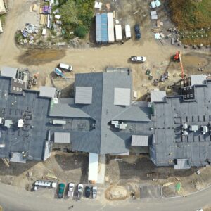A high up Ariel view from directly above a large building at the Piper Shores worksite. Around the building there are large containers, work vehicles, and construction materials.