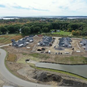An aerial view of a section of the paper stores community worksite. There are eight buildings under construction along with winding roads through the community and a large pond in the foreground. The coastline is visible in the background.