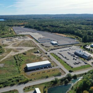 A high up aerial view of the Copart worksite. There is a large parking lot and three large buildings in the foreground, with the main worksite in the center. Large piles of gravel can be seen in the worksite as well as areas with gravel spread across them.