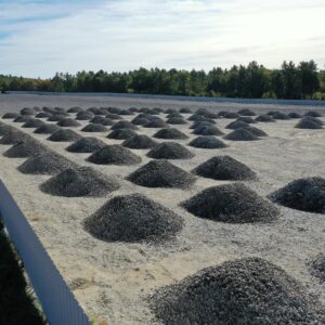 Many large piles of gravel evenly distributed across a fenced in area. Half of the area already has gravel spread evenly across it.