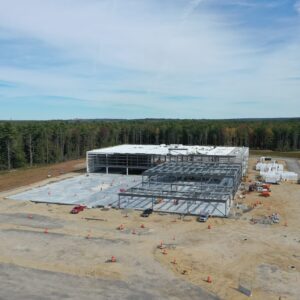 A high-up aerial view of a large building under construction at the IDXX worksite. A foundation slab is has been poured and there is a metal framework being constructed.
