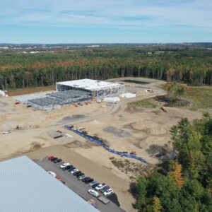 A high up aerial view of the IDEXX construction site with a large building under construction in the center and several piles of dirt and gravel around it. There is construction equipment around the worksite. The area is surrounded by trees.