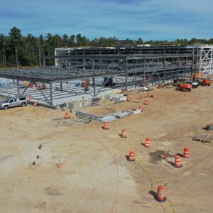 A large metal framework is being built for a building in the center of a dirt worksite. There are construction cones creating lanes for work trucks and lots of construction equipment is distributed across the worksite.