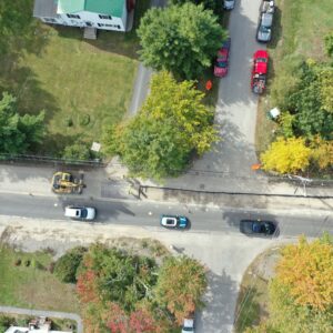 Aerial view of a road with the pavement removed from one lane, an excavator is parked near the end of the pavement and cars are driving in the other lane