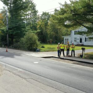 Road workers standing accross the street next to the area where pavement has been removed from the road